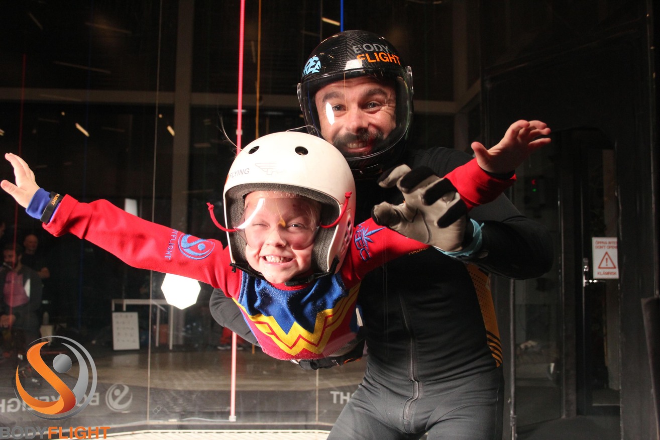 Liten tjej i wonderwomandräkt som testar på indoor skydiving bland sportlovsaktiviteter.